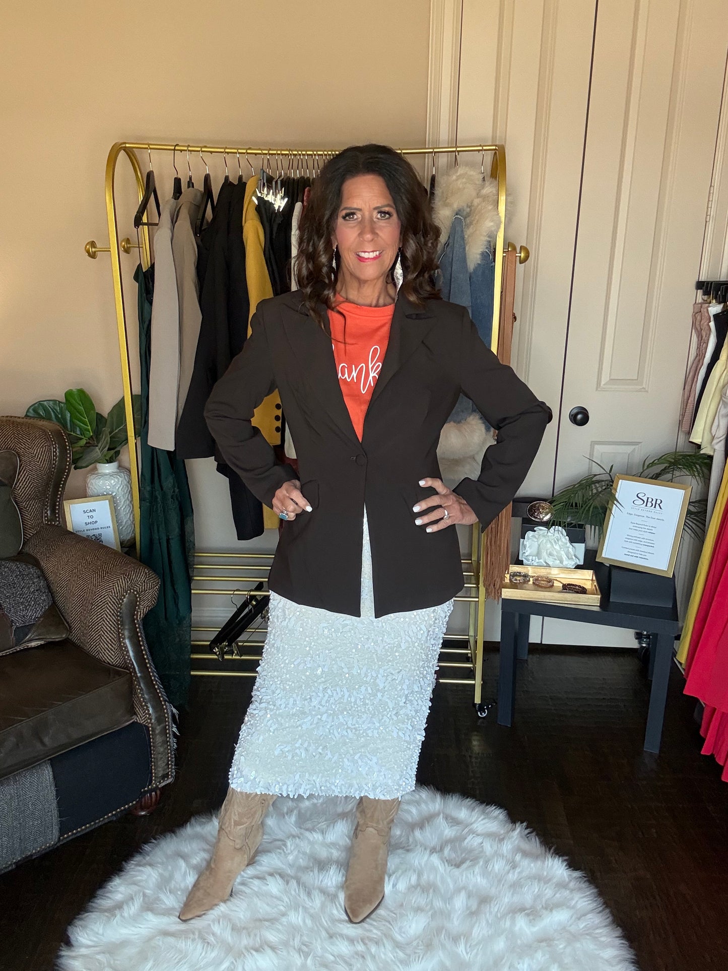 A woman stands in a room wearing a dark brown blazer over an orange graphic top and a white textured skirt, paired with beige boots. A clothing rack with various garments is visible behind her.