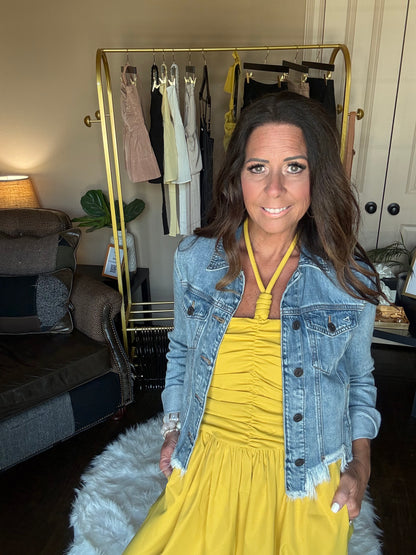 Woman wearing a yellow dress and denim jacket in a room with furniture and decor.