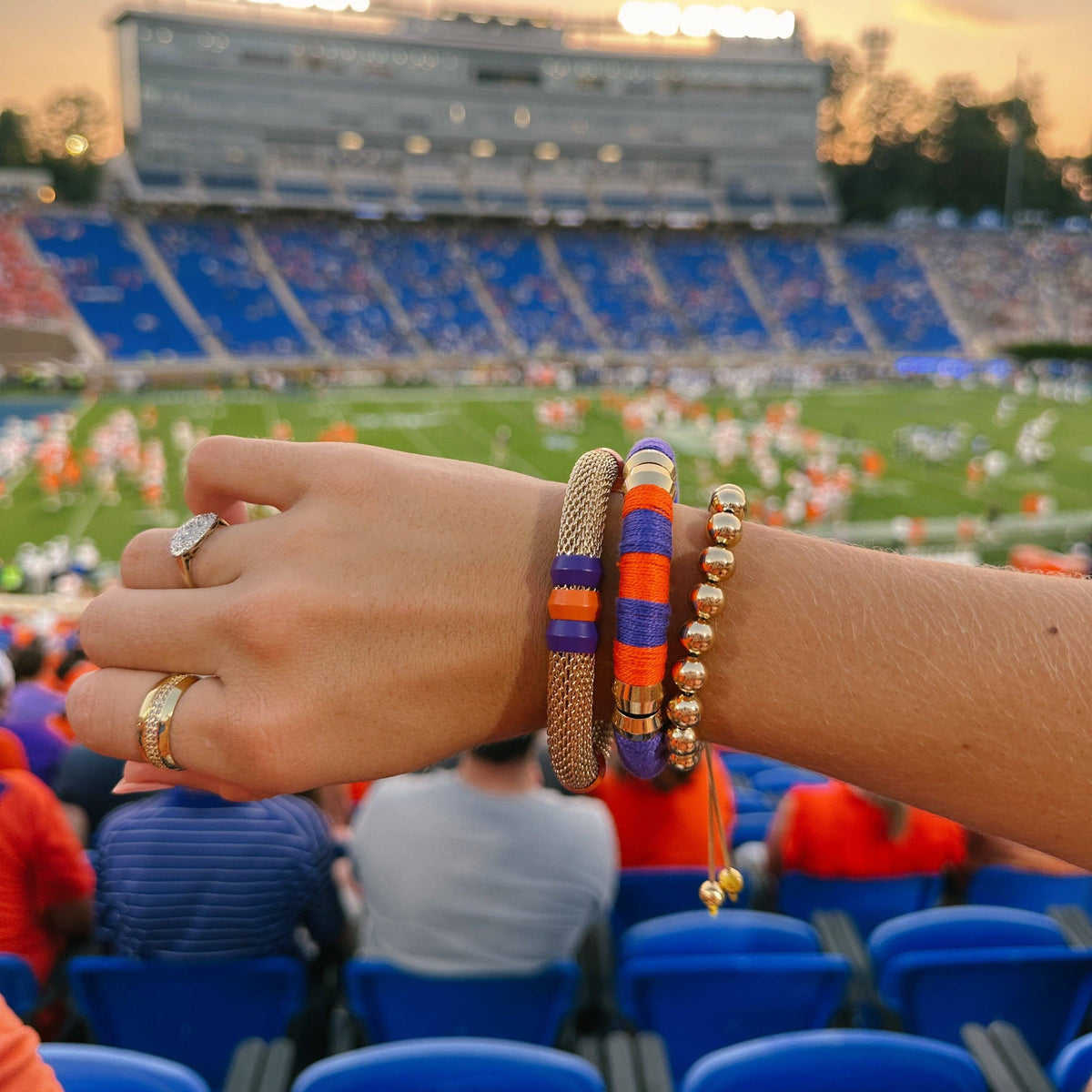 A close-up of a person's wrist wearing several bracelets at a football game, with stadium seating and players visible in the background. The bracelets include a woven tan bangle, a blue and orange striped bangle, a purple and orange striped bangle, and a gold beaded bangle.