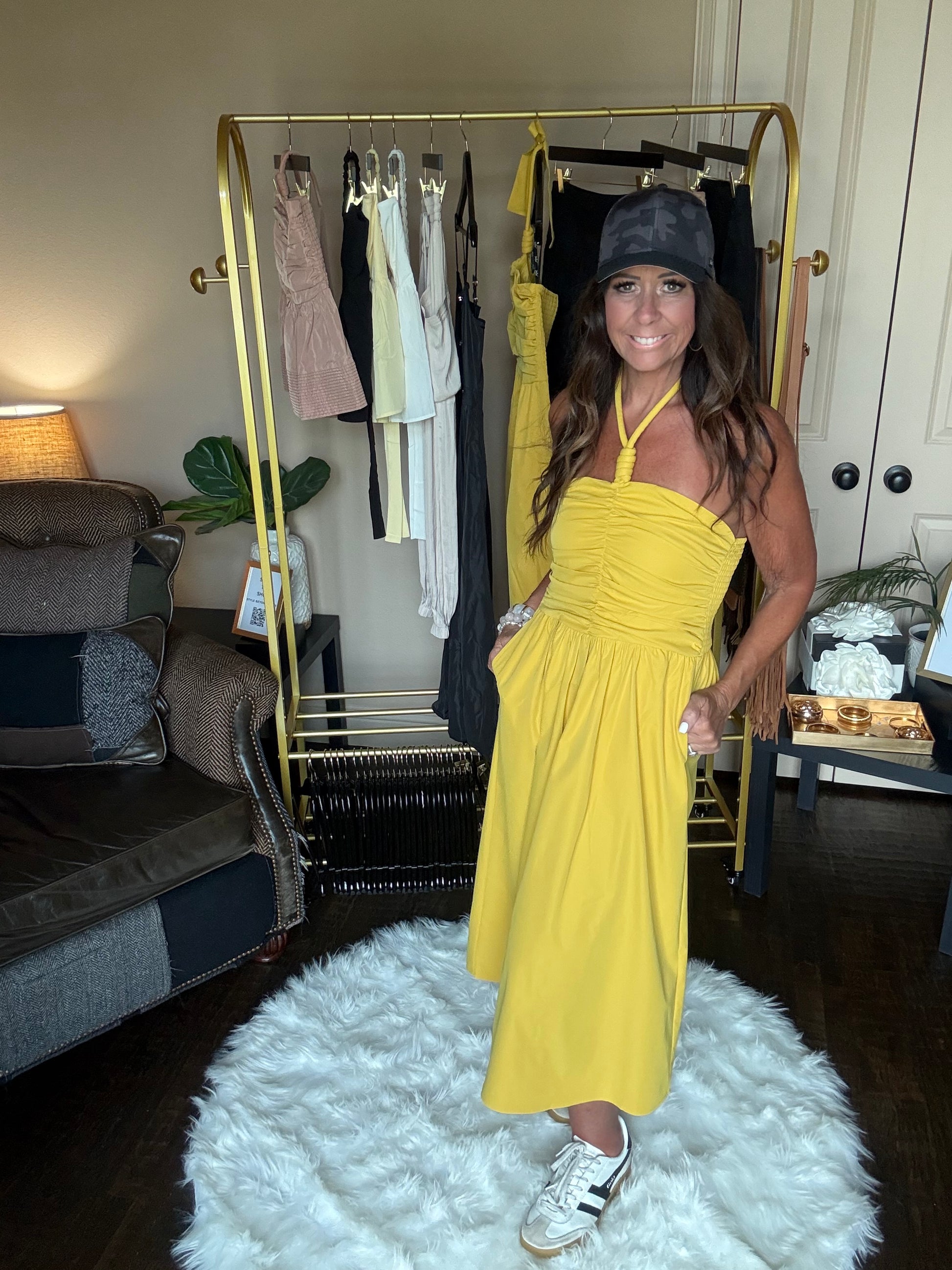 Woman in a yellow dress standing in front of a gold mirror in a room with a white rug.