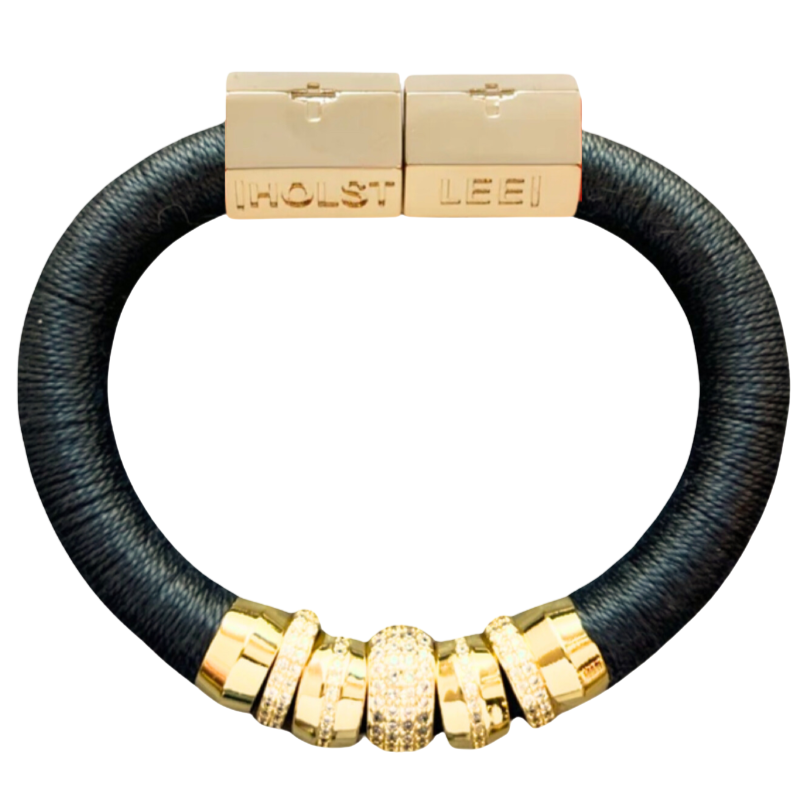Holst + Lee charcoal braided cord bracelet with gold-toned hardware, featuring two rectangular end caps engraved with 'HOLST' and 'LEE', and a row of gold spacer beads with clear crystal detailing near the bottom.
