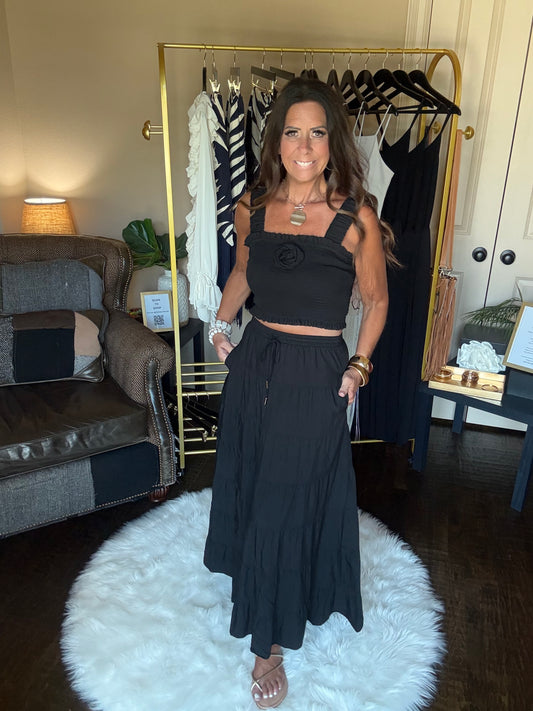 A woman modeling a black two-piece outfit consisting of a sleeveless crop top with a flower detail and a matching long skirt, standing in a room with a clothing rack and a white fluffy rug.