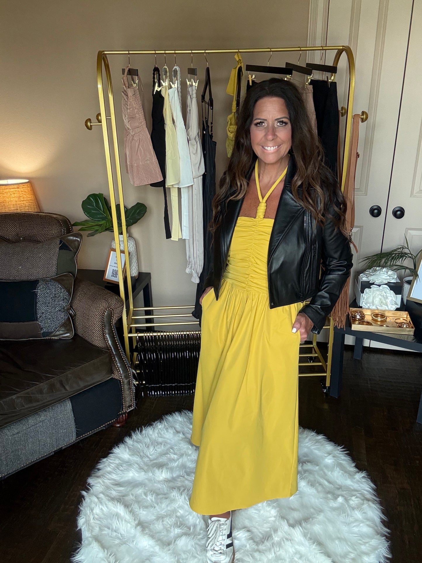 Woman in a yellow dress and black leather jacket standing in a room with a gold bar cart and white rug.