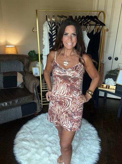 A woman wearing a brown and beige zebra-print mini dress, posing in front of a gold clothing rack inside a home setting.