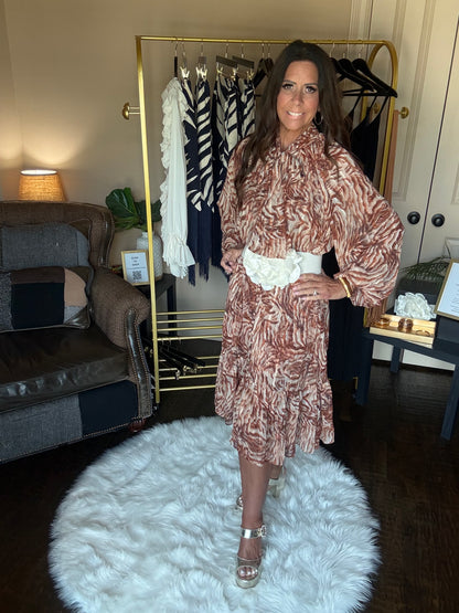 A woman modeling a brown and beige patterned long-sleeve dress with a white lace detail at the waist, standing in a room with a clothing rack, sofa, and a white fluffy rug.