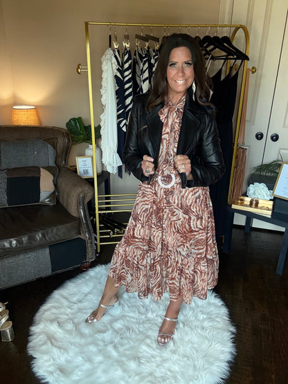 A woman wearing a brown and beige patterned midi dress with a black leather jacket and holding a black bag, standing in a room with a gold clothing rack, a white shag rug, and a couch.