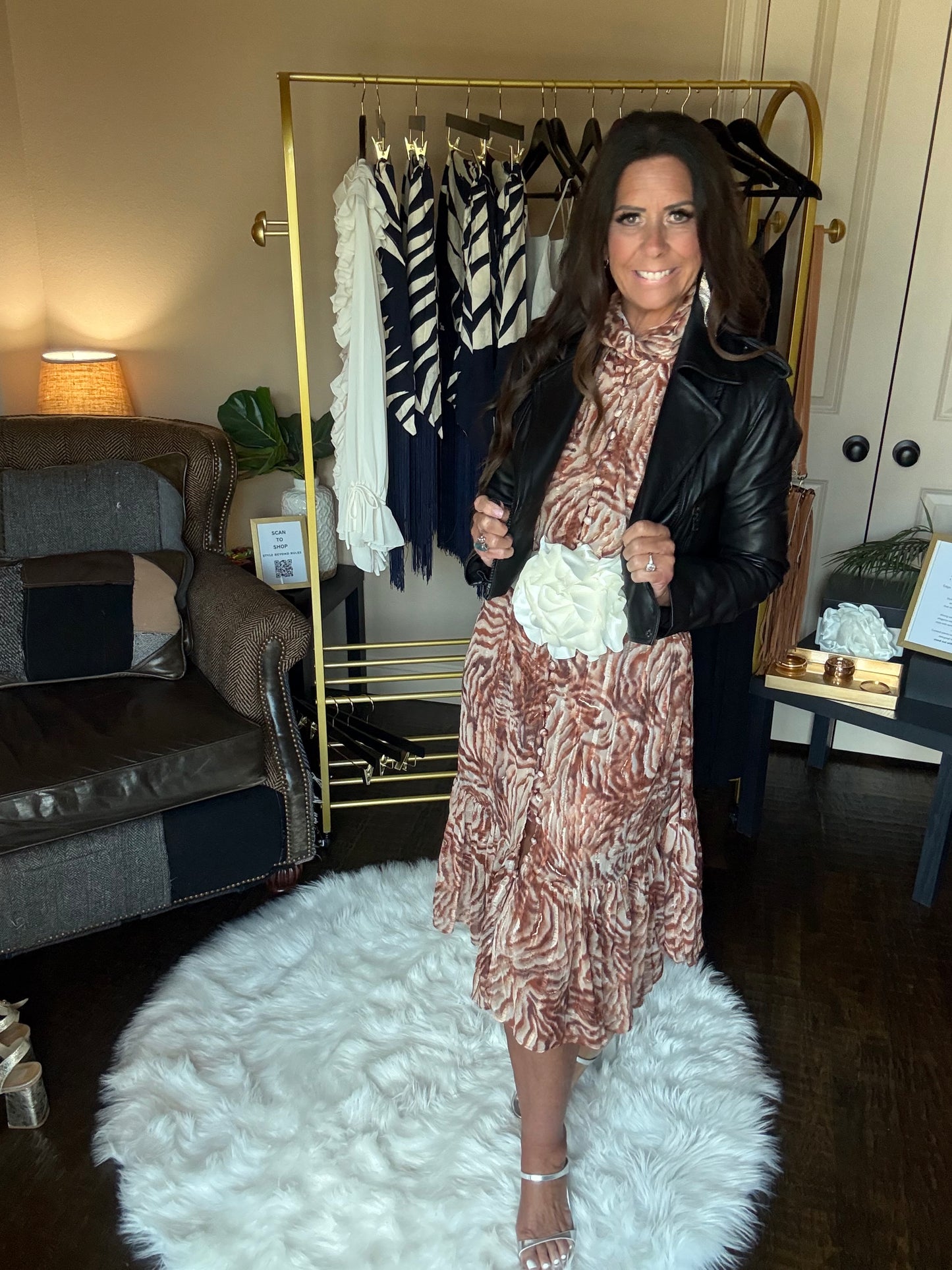 A woman wearing a brown and beige patterned dress with a black leather jacket, standing in a room with a gold clothing rack, a white fluffy rug, and a couch.