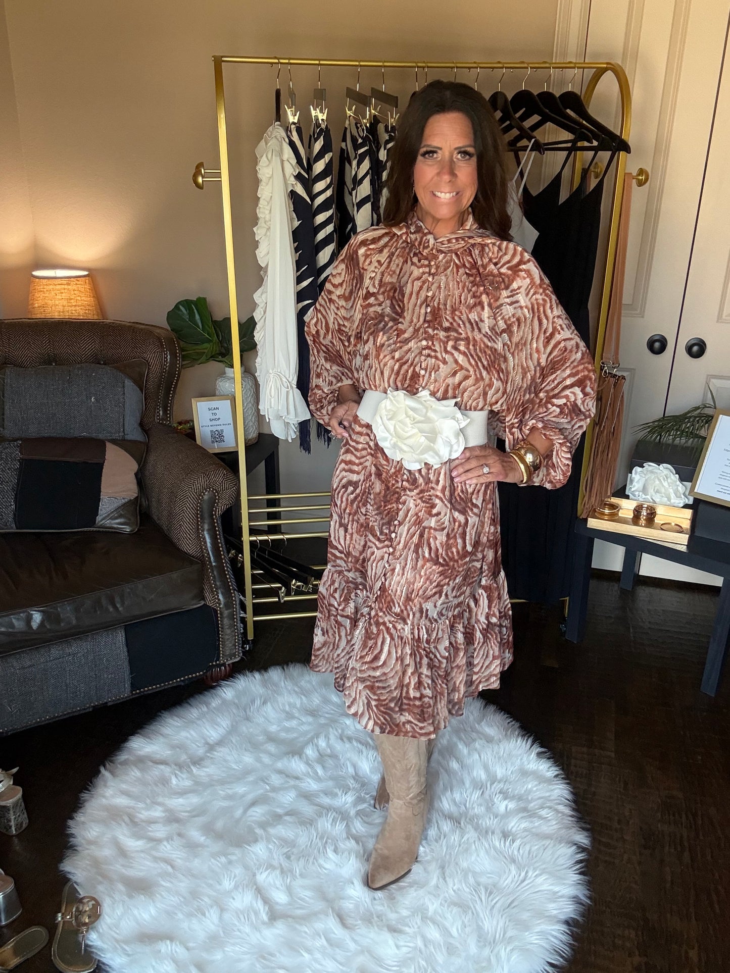 A woman wearing a brown and beige zebra-print dress with a white floral accessory, standing in a room with a gold clothing rack, a white shag rug, and a sofa.
