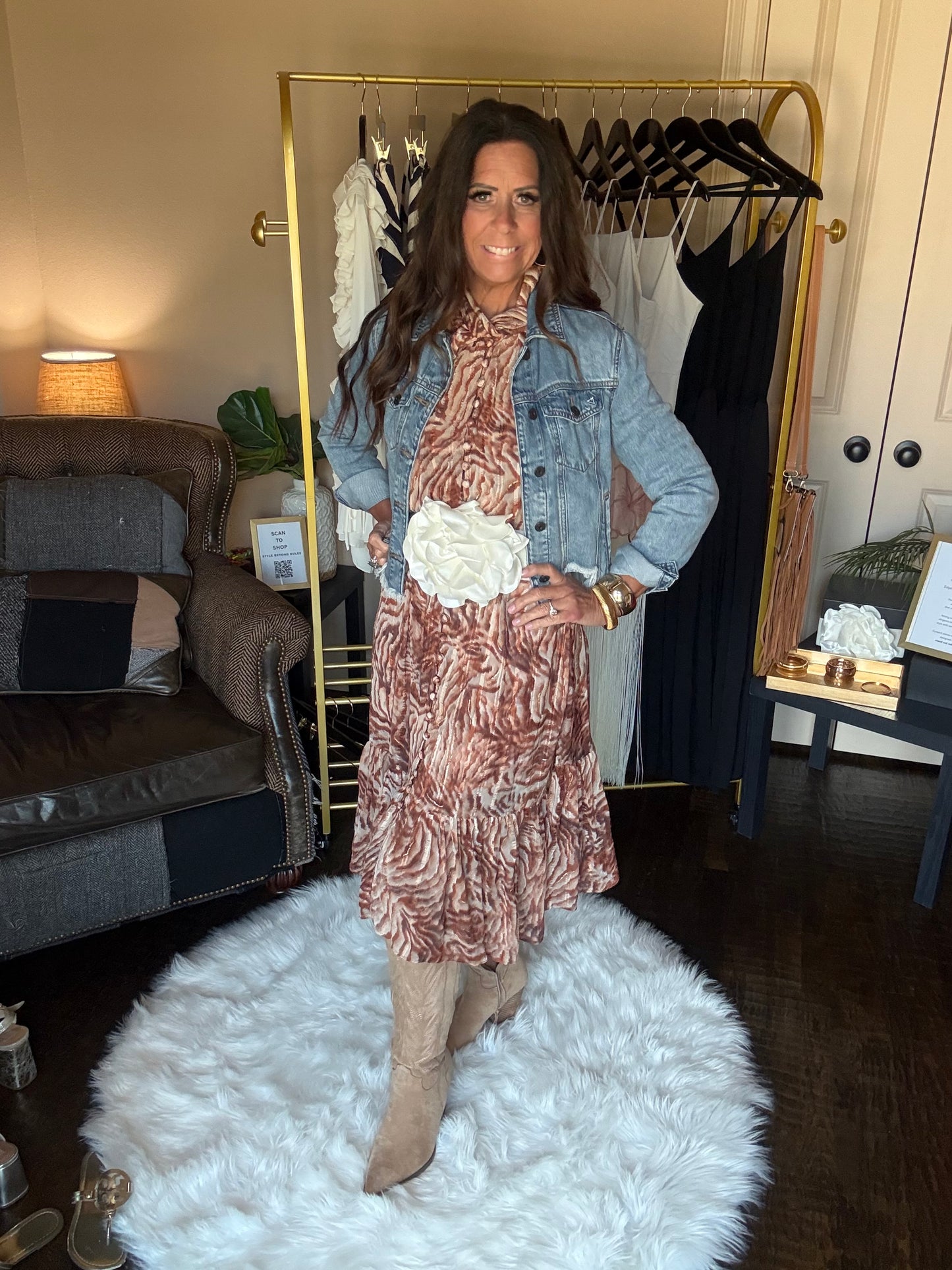 A woman models a patterned brown and pink dress with a light blue denim jacket and a white flower accessory, standing in a room with a gold-framed mirror, clothing rack, and a white faux fur rug.