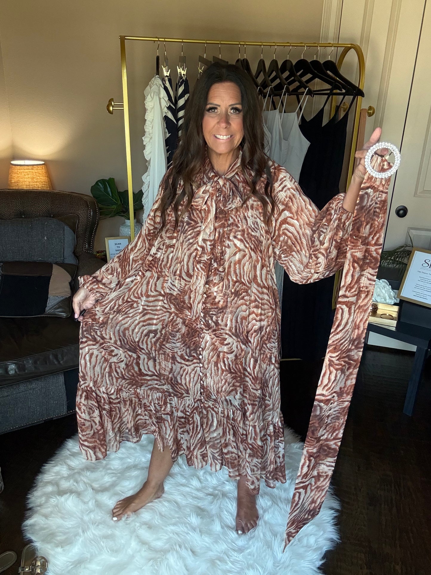 A woman wearing a brown and beige zebra-print kaftan dress, holding up the sleeves of the dress in a home setting with a white faux fur rug and a clothing rack in the background.