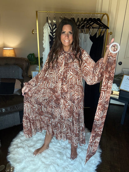 A woman wearing a brown and beige zebra-print kaftan dress, holding up the sleeves of the dress in a home setting with a white faux fur rug and a clothing rack in the background.