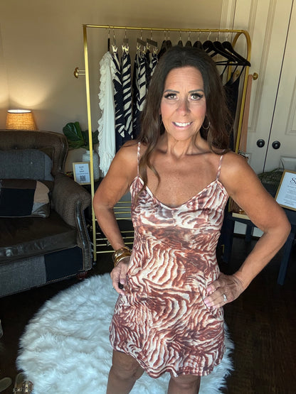 A woman wearing a brown and beige zebra-print spaghetti-strap dress, standing in a home setting with a clothing rack and sofa in the background.