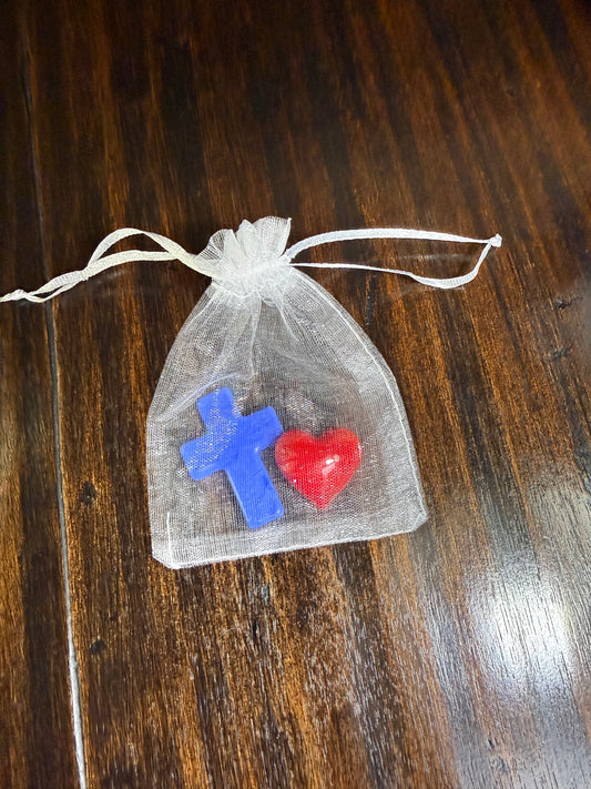 A small white organza drawstring pouch containing a red heart-shaped charm and a blue cross-shaped charm, placed on a dark wooden surface.