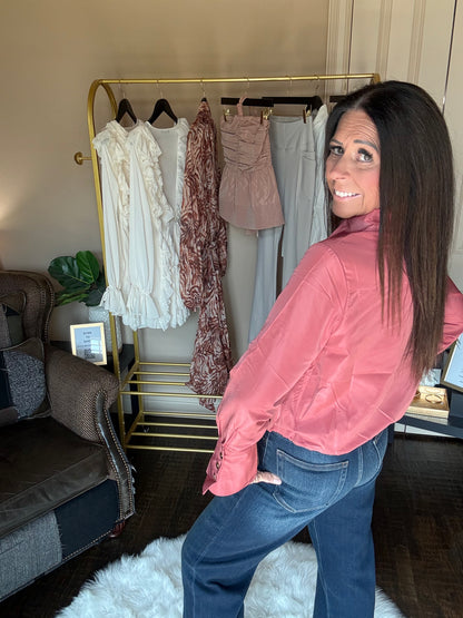 A woman with long dark hair stands in a room wearing a shiny pink satin blouse and blue jeans, looking back over her shoulder. A gold clothing rack with several garments is behind her, along with a chair, plant, and rug.
