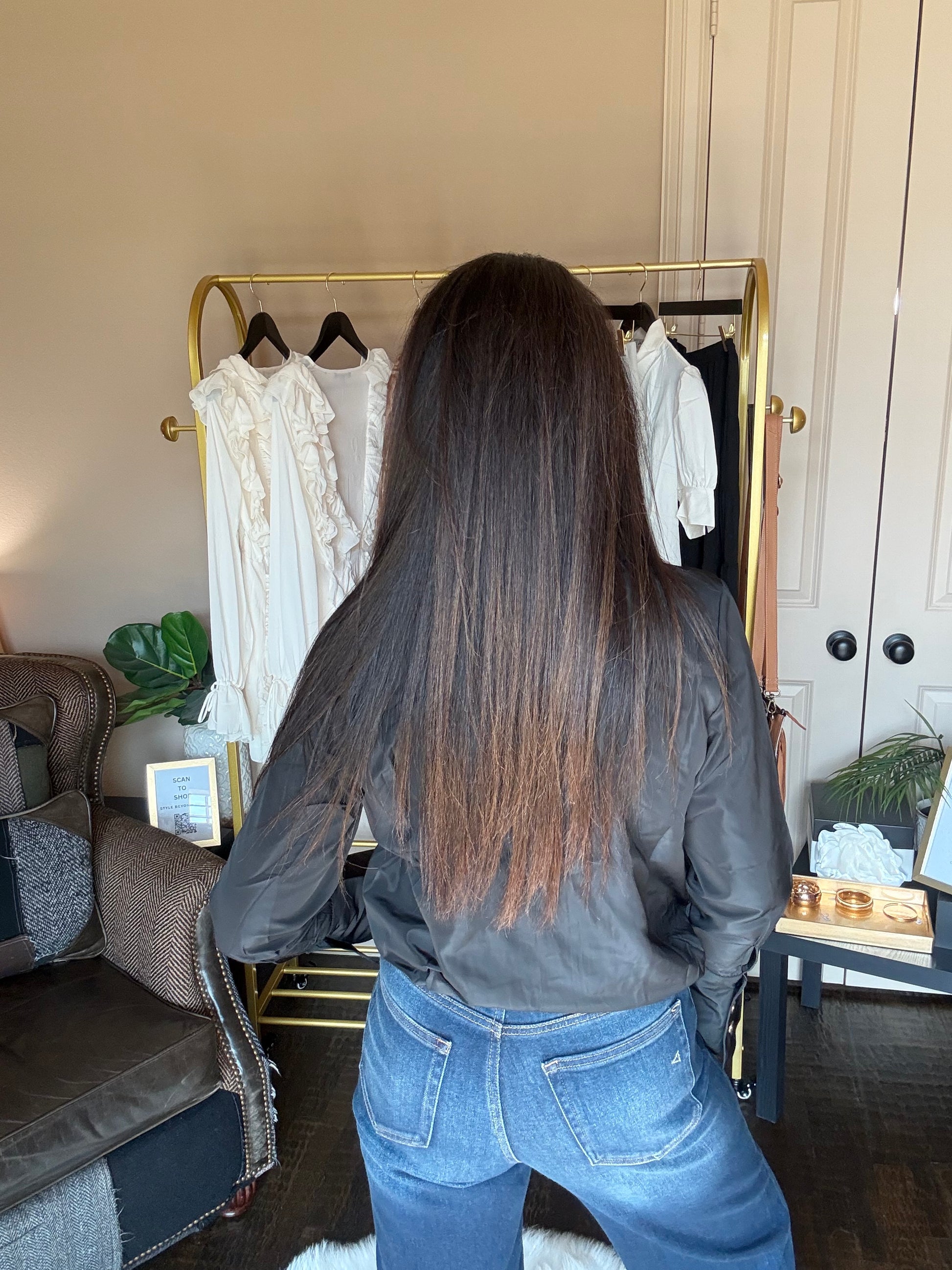 A person with very long, straight dark brown hair stands with their back to the camera in a home setting, wearing a black leather jacket and blue jeans. A gold clothing rack with white and black tops is visible behind them.