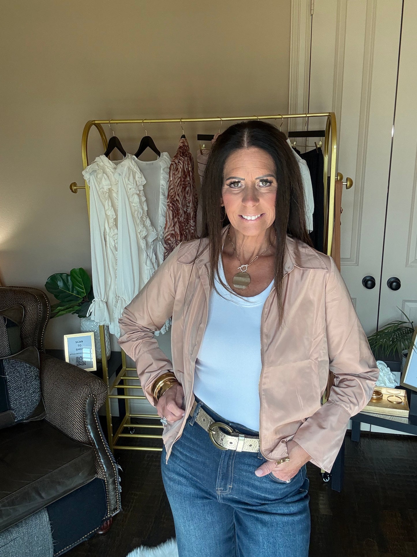 A woman stands indoors wearing a light beige satin-like jacket over a white tank top and blue jeans, with a gold belt. A clothing rack with white and rust-colored garments is behind her, along with a chair and room interior.