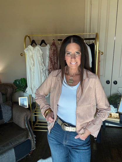 A woman stands indoors wearing a light beige satin-like jacket over a white tank top and blue jeans, with a gold belt. A clothing rack with white and rust-colored garments is behind her, along with a chair and room interior.
