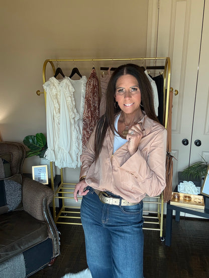 A woman stands in a room wearing a light beige button-up shirt and blue jeans, standing in front of a gold clothing rack with several shirts hanging on it.