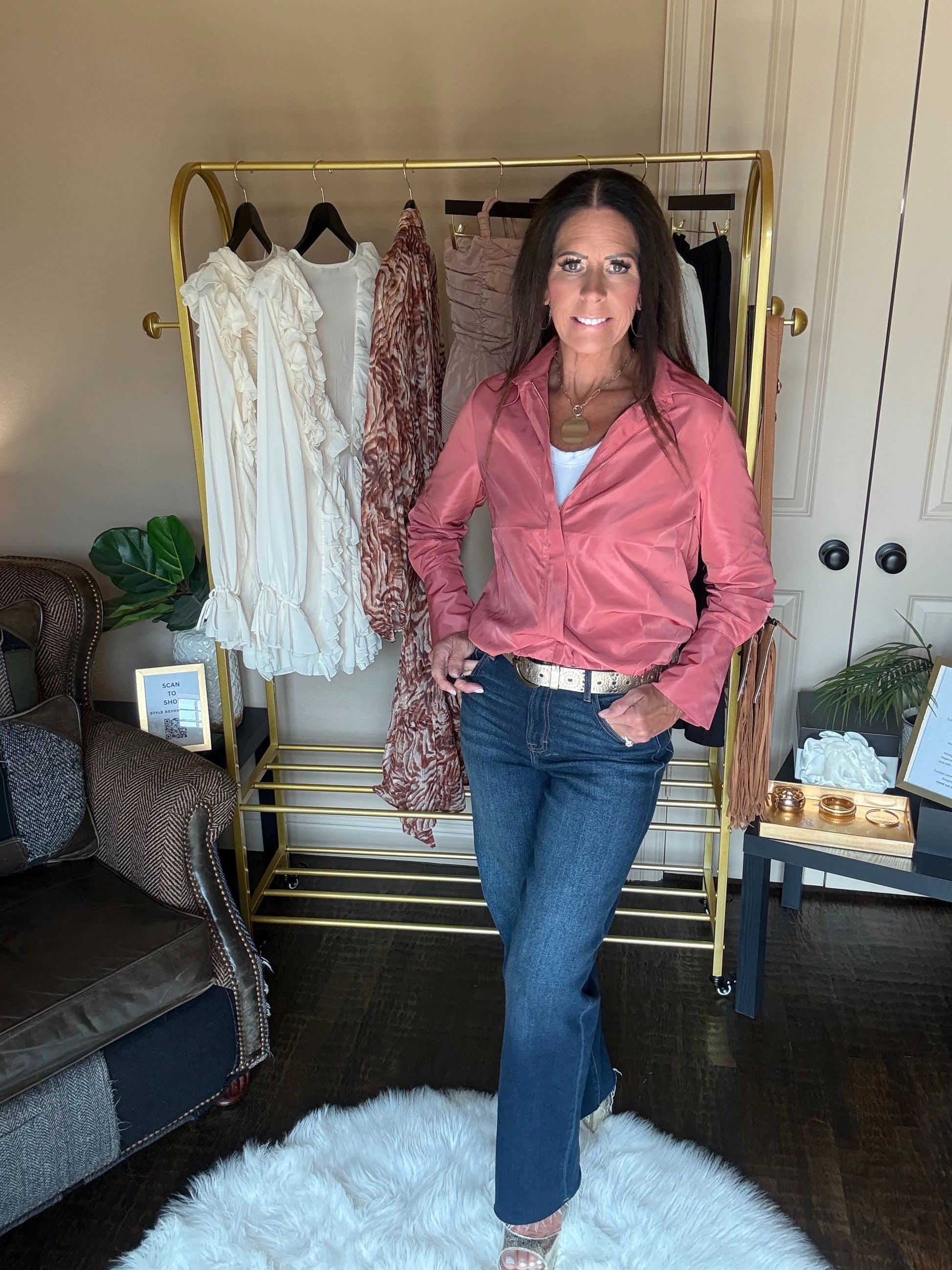 A woman stands indoors wearing a dusty pink satin button-up shirt and blue jeans, posing in front of a gold clothing rack filled with other garments.