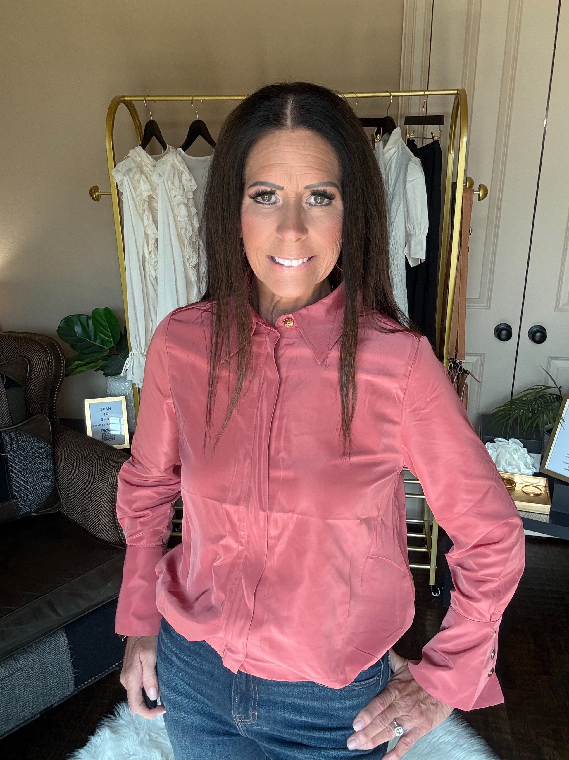 A woman with long dark hair wearing a dusty rose pink satin long-sleeve blouse and blue jeans, standing in front of a gold clothing rack in a home setting.