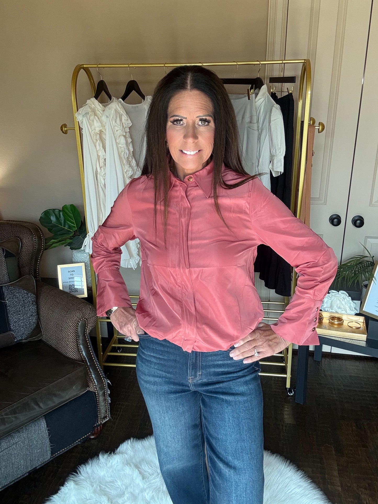 A woman wearing a dusty rose pink long-sleeve zip-up jacket and blue jeans stands in front of a gold clothing rack in a home setting.