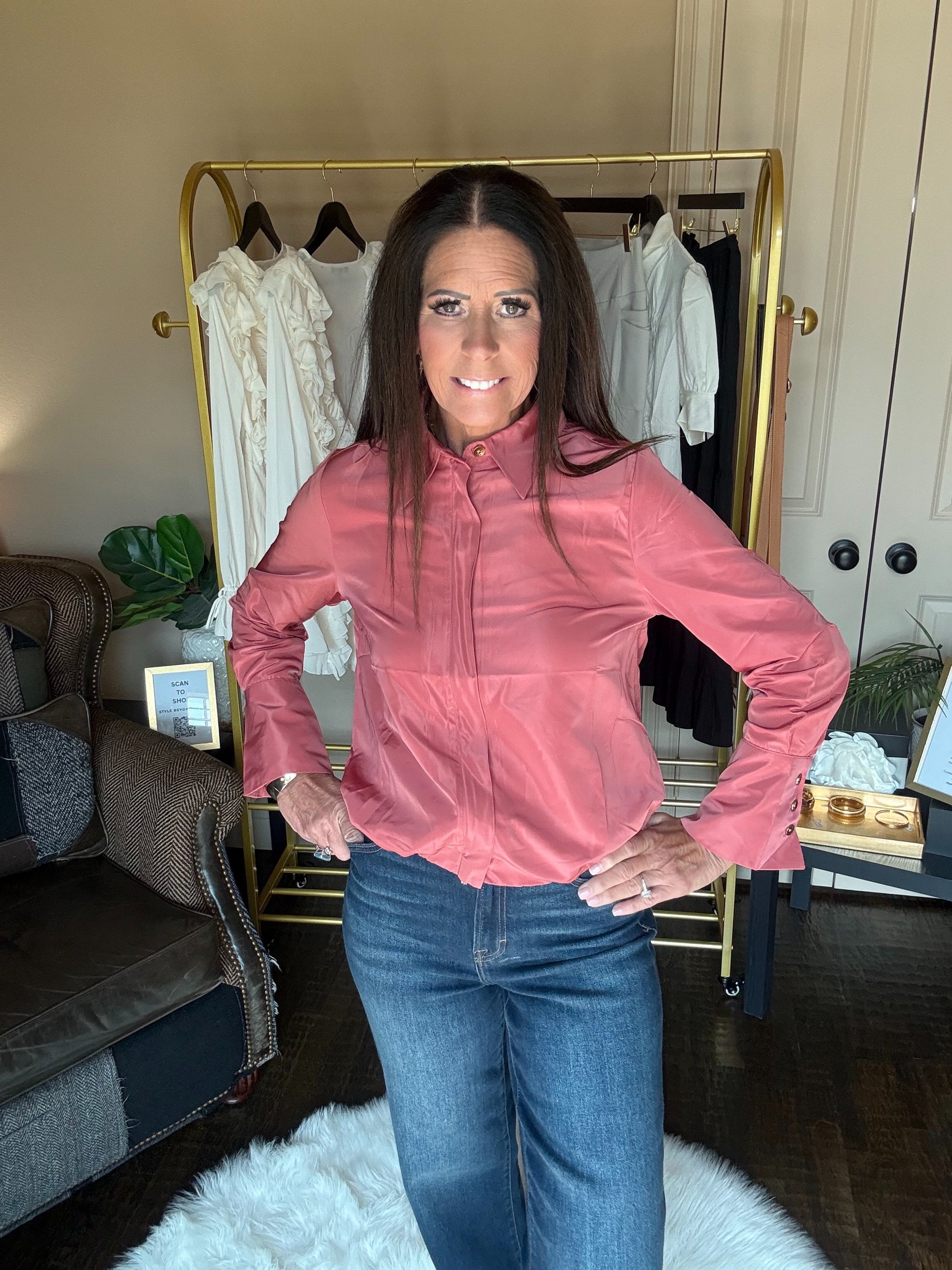 A woman wearing a dusty rose pink long-sleeve zip-up jacket and blue jeans stands in front of a gold clothing rack in a home setting.