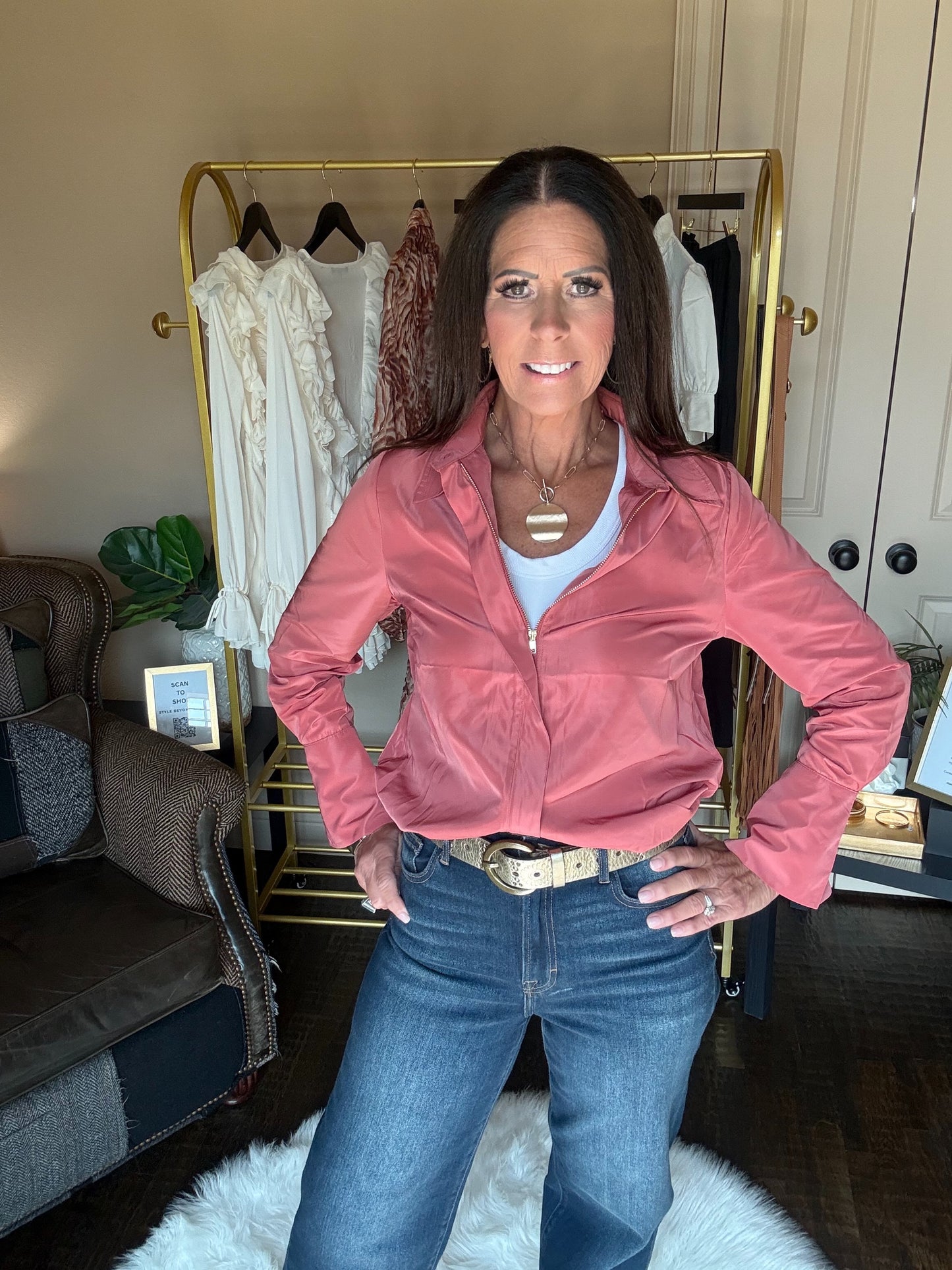 A woman posing in a dusty pink zip-up jacket, blue jeans, and a white tank top in a home setting with a gold clothing rack behind her.