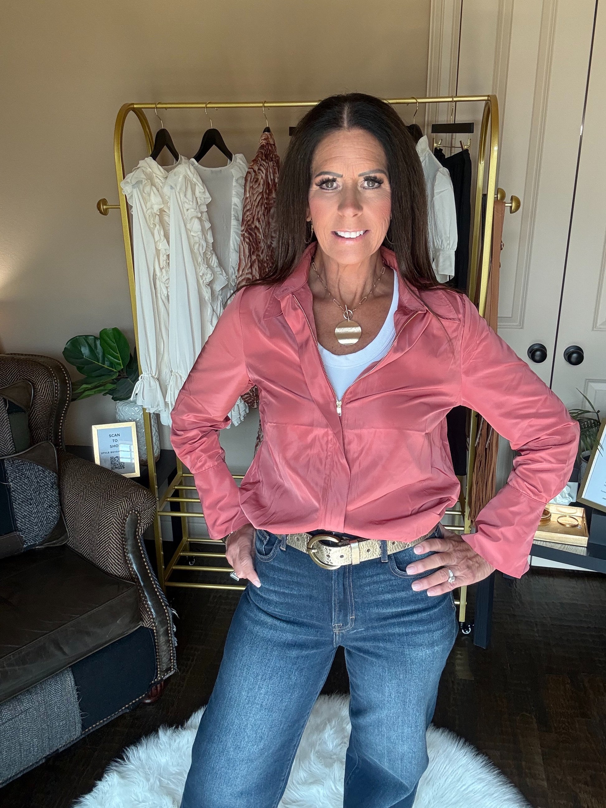 A woman posing in a dusty pink zip-up jacket, blue jeans, and a white tank top in a home setting with a gold clothing rack behind her.