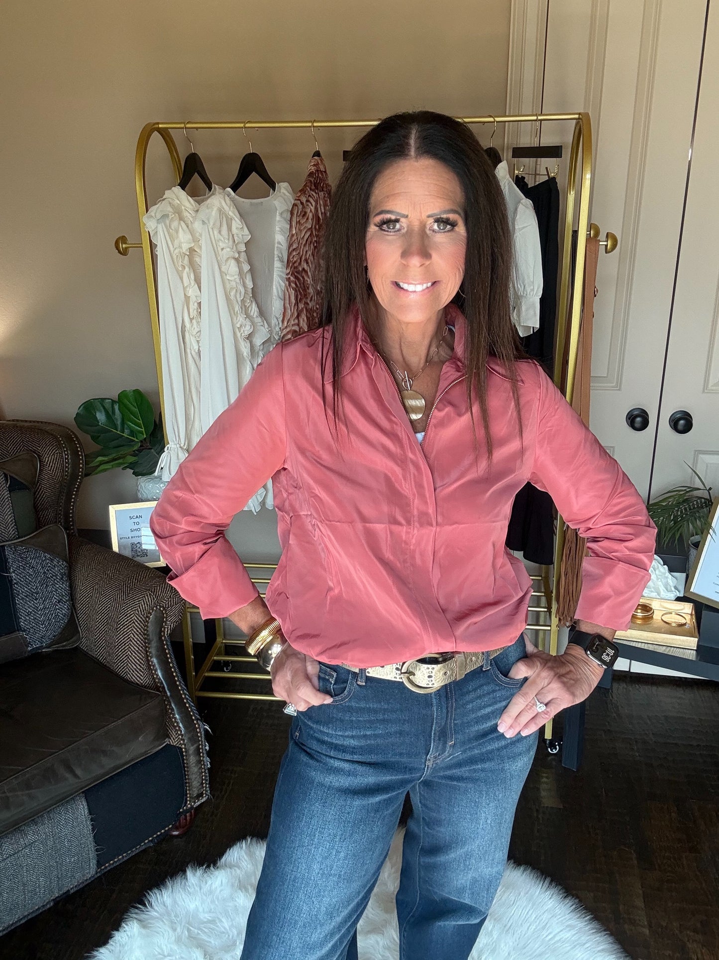 A woman wearing a dusty pink button-down shirt and blue jeans stands with hands on hips in front of a gold clothing rack in a home setting.