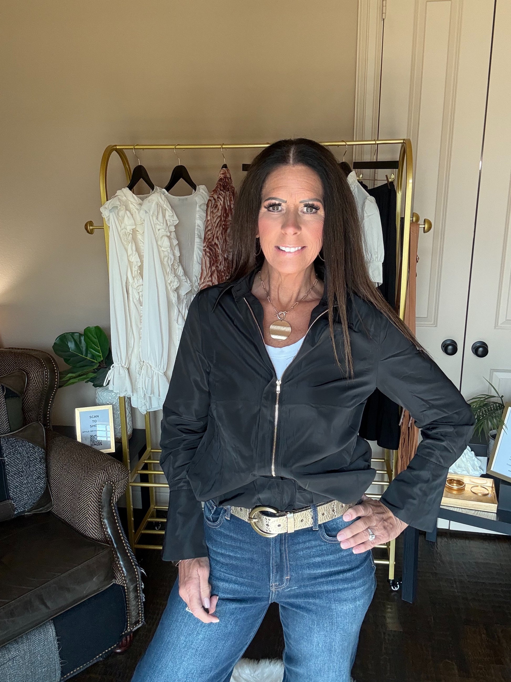 A woman modeling a black zip-up jacket with a gold zipper, worn over a white top and paired with blue jeans, standing in front of a clothing rack in an indoor setting.