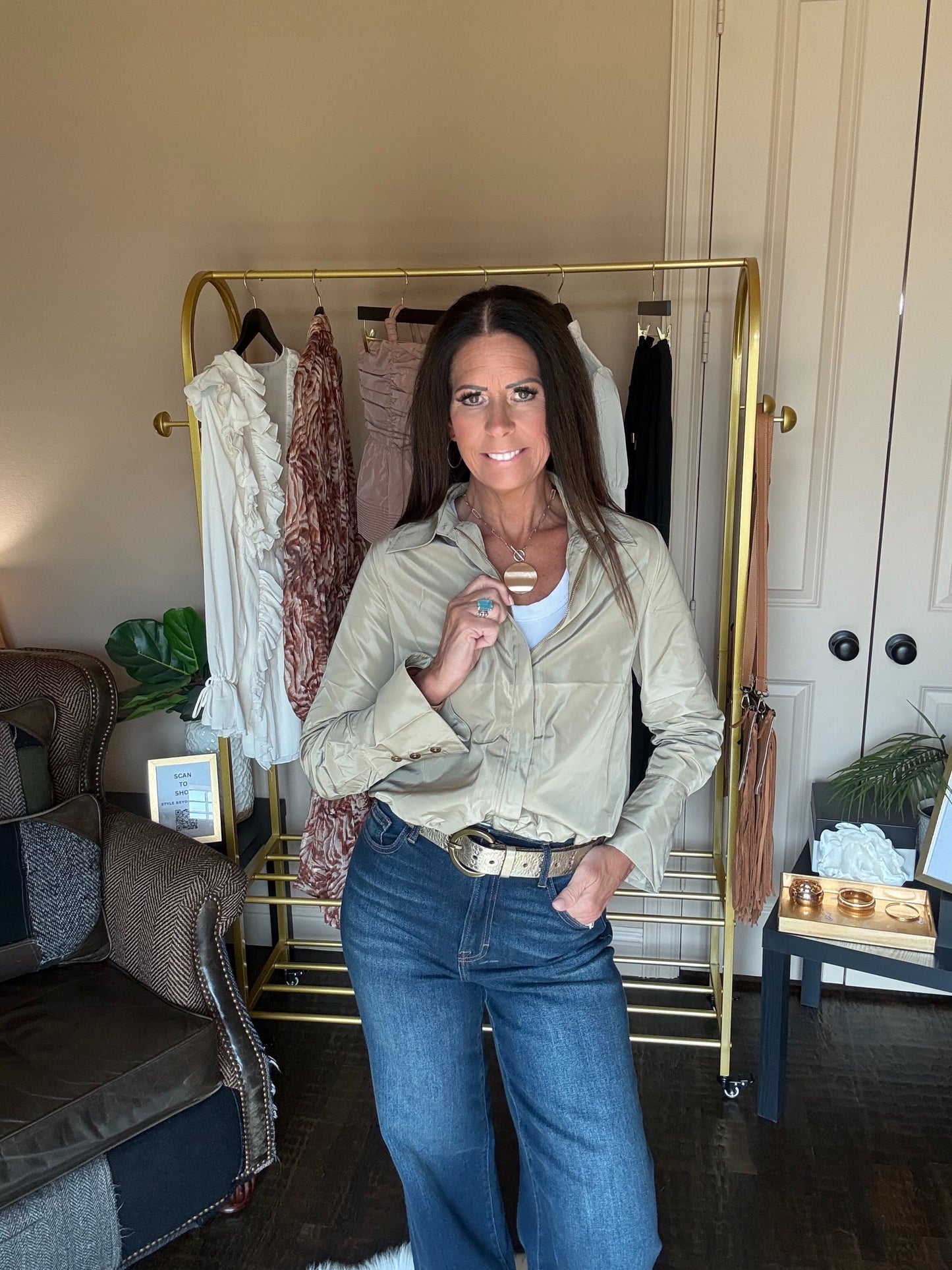 A woman stands indoors wearing a light beige button-up shirt and blue jeans, with a gold clothing rack and furniture visible in the background.