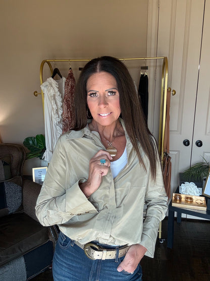 A woman with long dark hair stands indoors wearing a light beige button-up shirt over a white top and blue jeans, with a gold belt. A clothing rack with garments is visible behind her in a home setting.