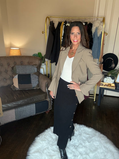 Model is wearing a khaki blazer with a cream top, black ribbed maxi skirt with gold button details, and black booties