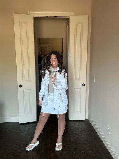 Woman standing in a room wearing a light-colored trench coat.