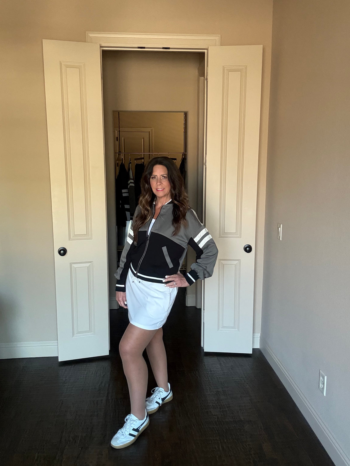Woman standing in a room with open doors, wearing a black and gray jacket and white shorts.
