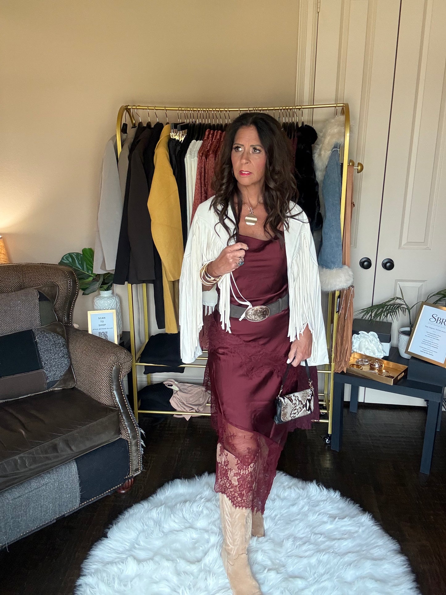 A woman stands in a room wearing a burgundy satin midi dress with lace hem, a white fringed cardigan, beige boots, and holding a small metallic handbag. A clothing rack, sofa, and side table are visible in the background.