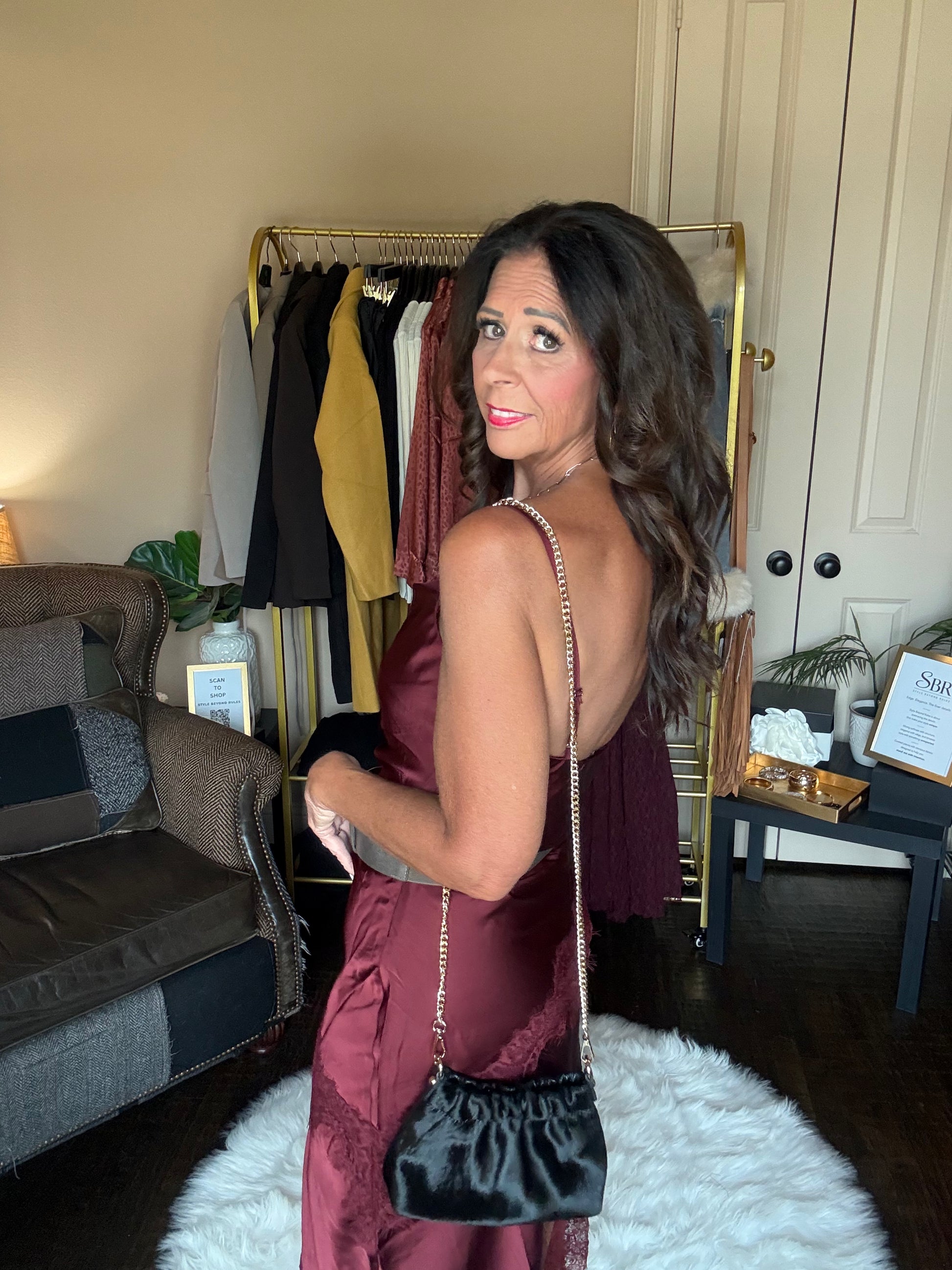 A woman wearing a backless burgundy satin dress and carrying a black handbag with a silver chain strap, standing in a room with a clothing rack and furniture in the background.