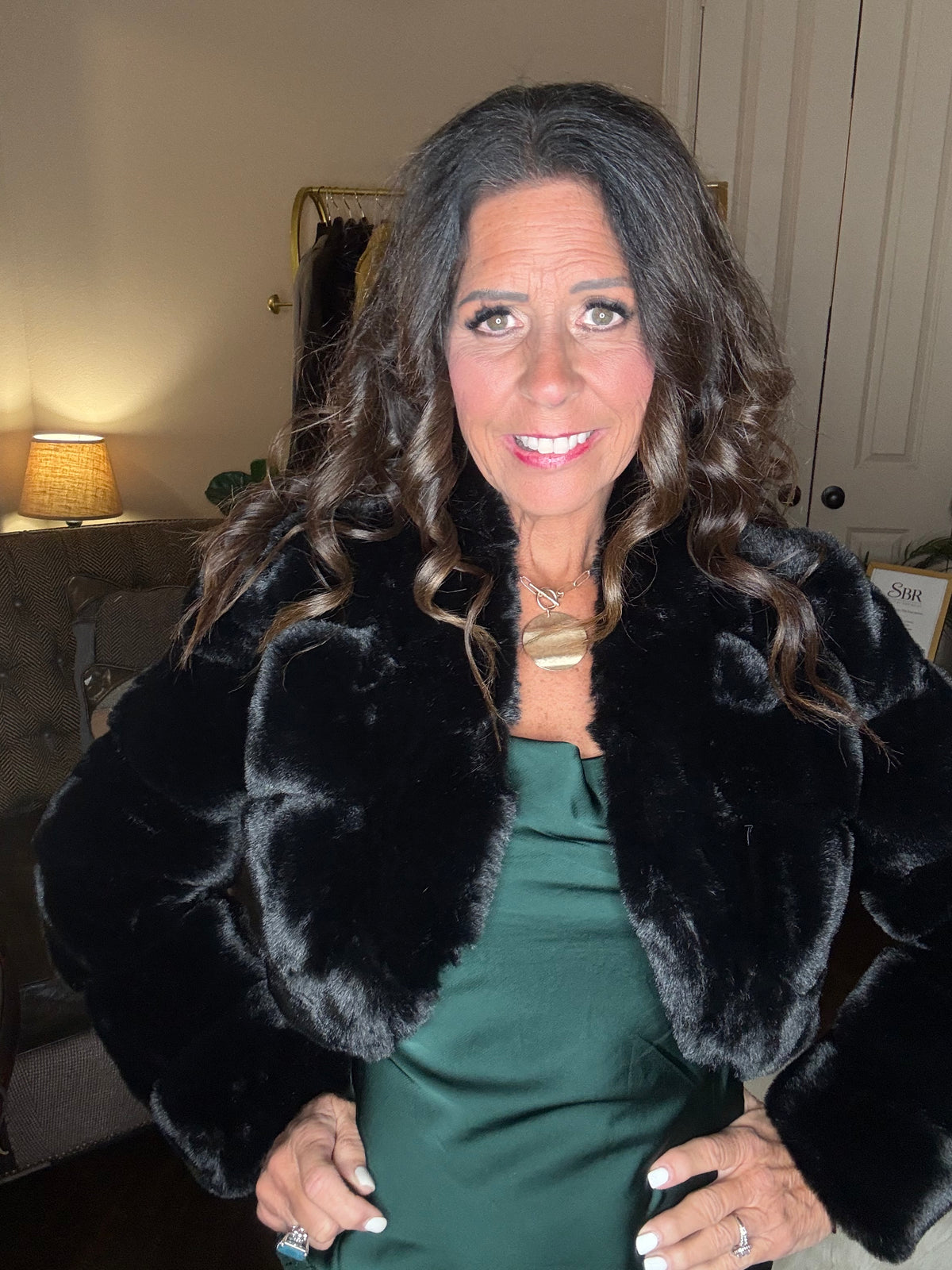 Model is wearing a hunter green satin and lace maxi dress with spaghetti straps and an asymmetrical hem layered with a black faux fur bolero jacket.