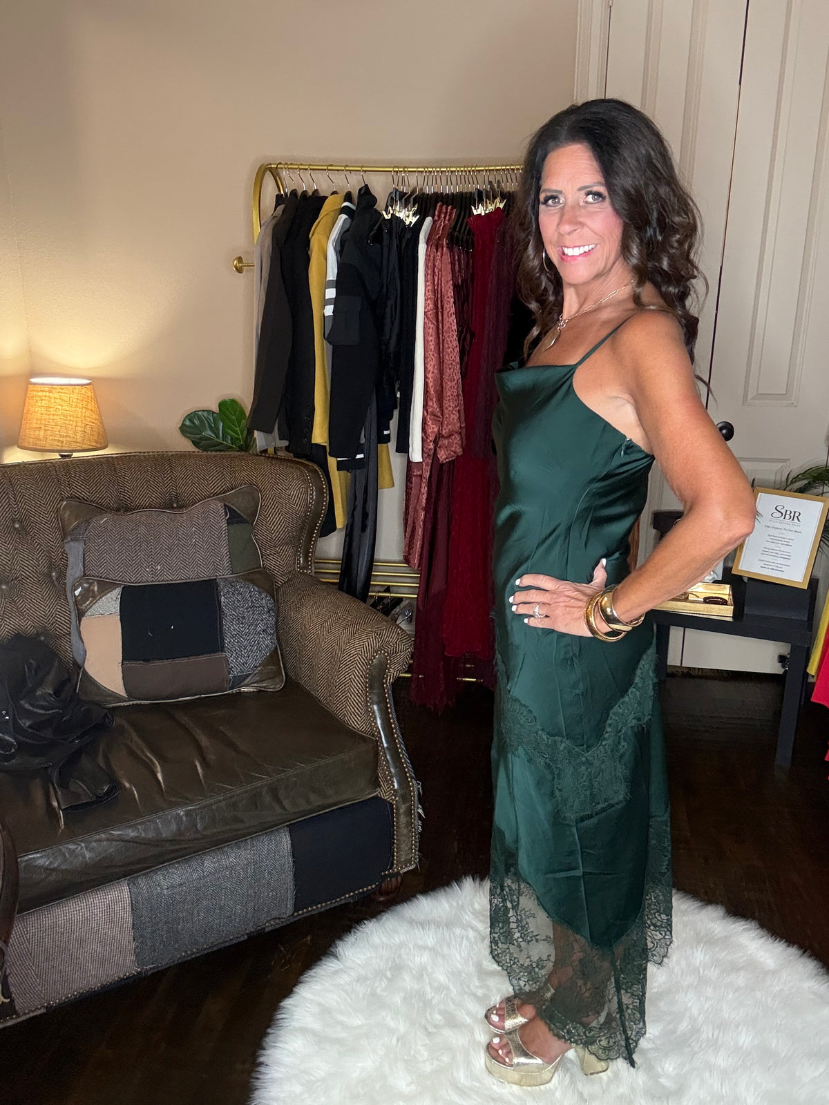 Model is wearing a hunter green satin and lace maxi dress with spaghetti straps and an asymmetrical hem land gold metallic heels.
