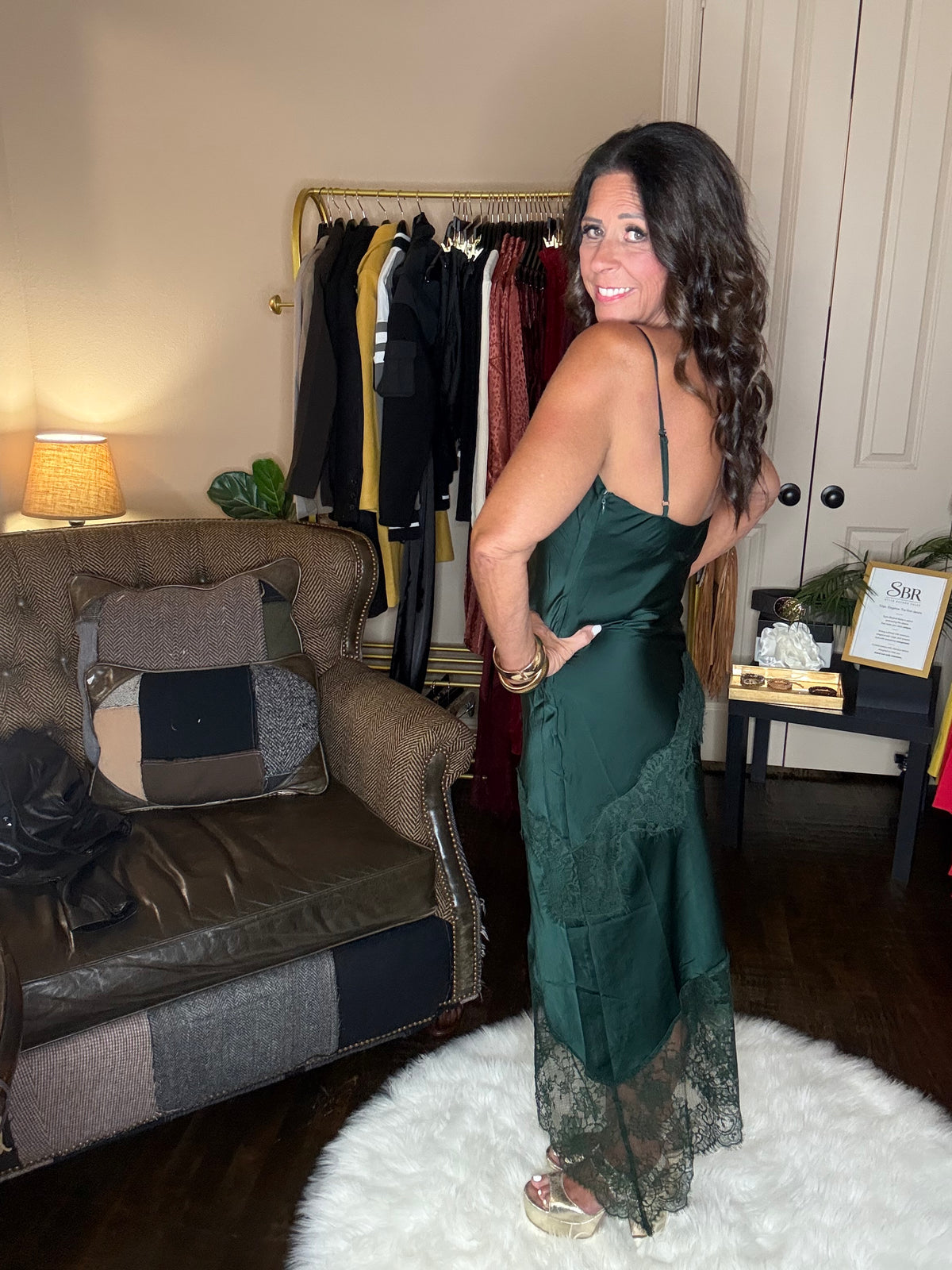 Model is wearing a hunter green satin and lace maxi dress with spaghetti straps and an asymmetrical hem land gold metallic heels.