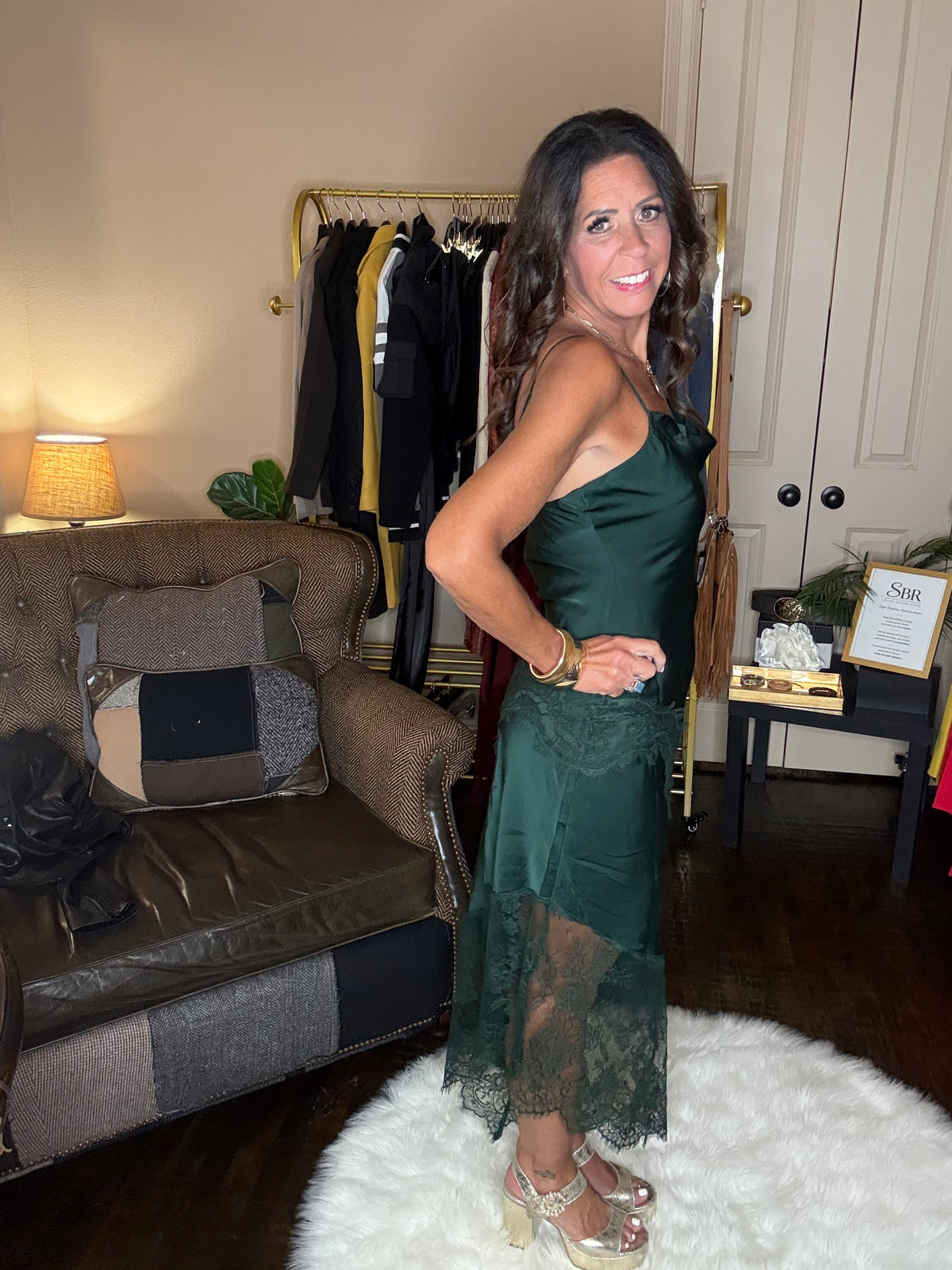 Model is wearing a hunter green satin and lace maxi dress with spaghetti straps and an asymmetrical hem land gold metallic heels.