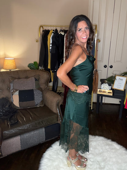 Model is wearing a hunter green satin and lace maxi dress with spaghetti straps and an asymmetrical hem land gold metallic heels.