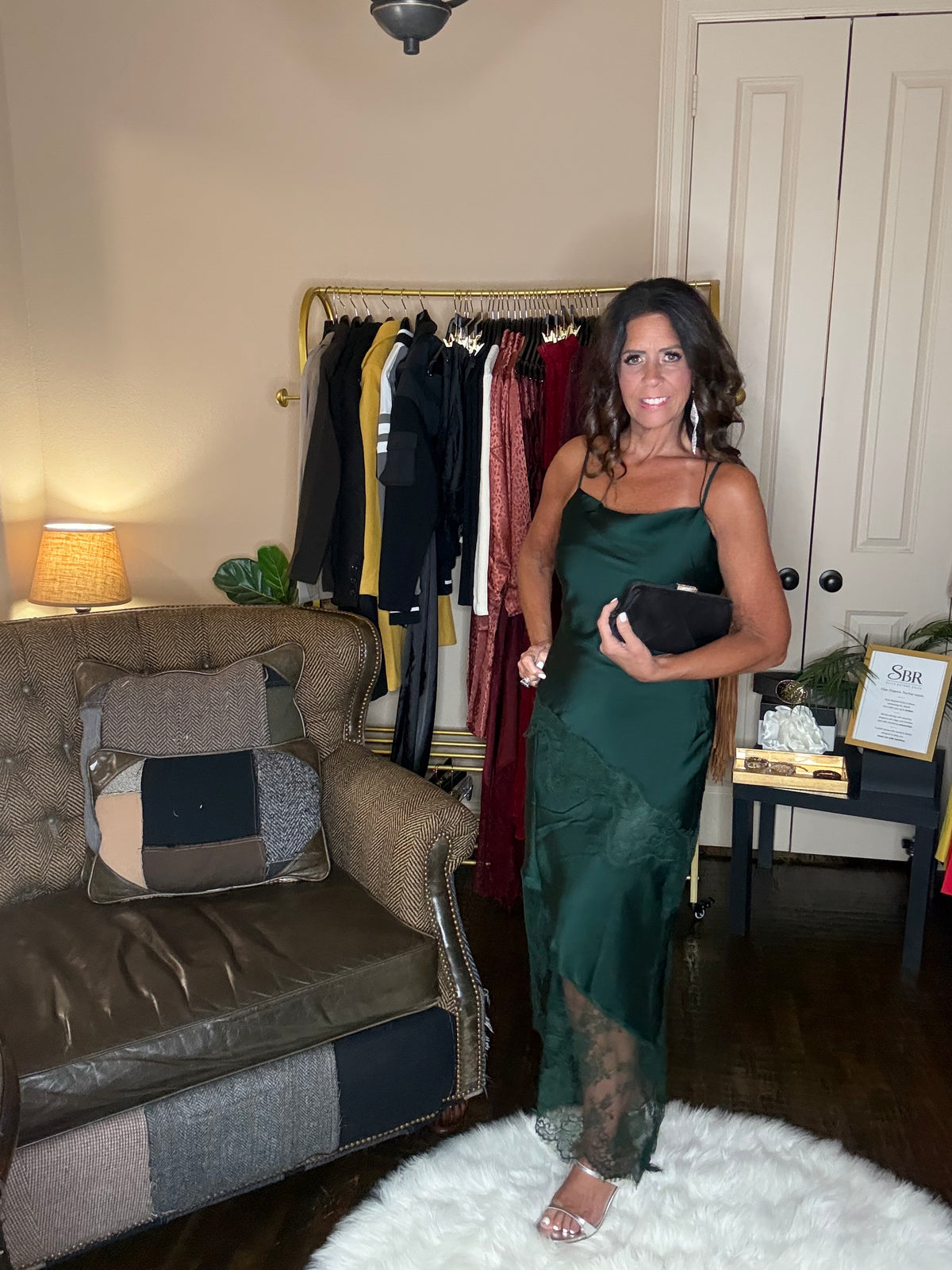 Model is wearing a hunter green satin and lace maxi dress with spaghetti straps and an asymmetrical hem land gsilver metallic heels.