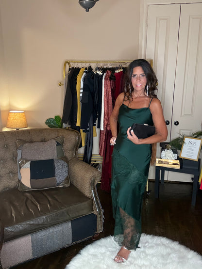 Model is wearing a hunter green satin and lace maxi dress with spaghetti straps and an asymmetrical hem land gsilver metallic heels.