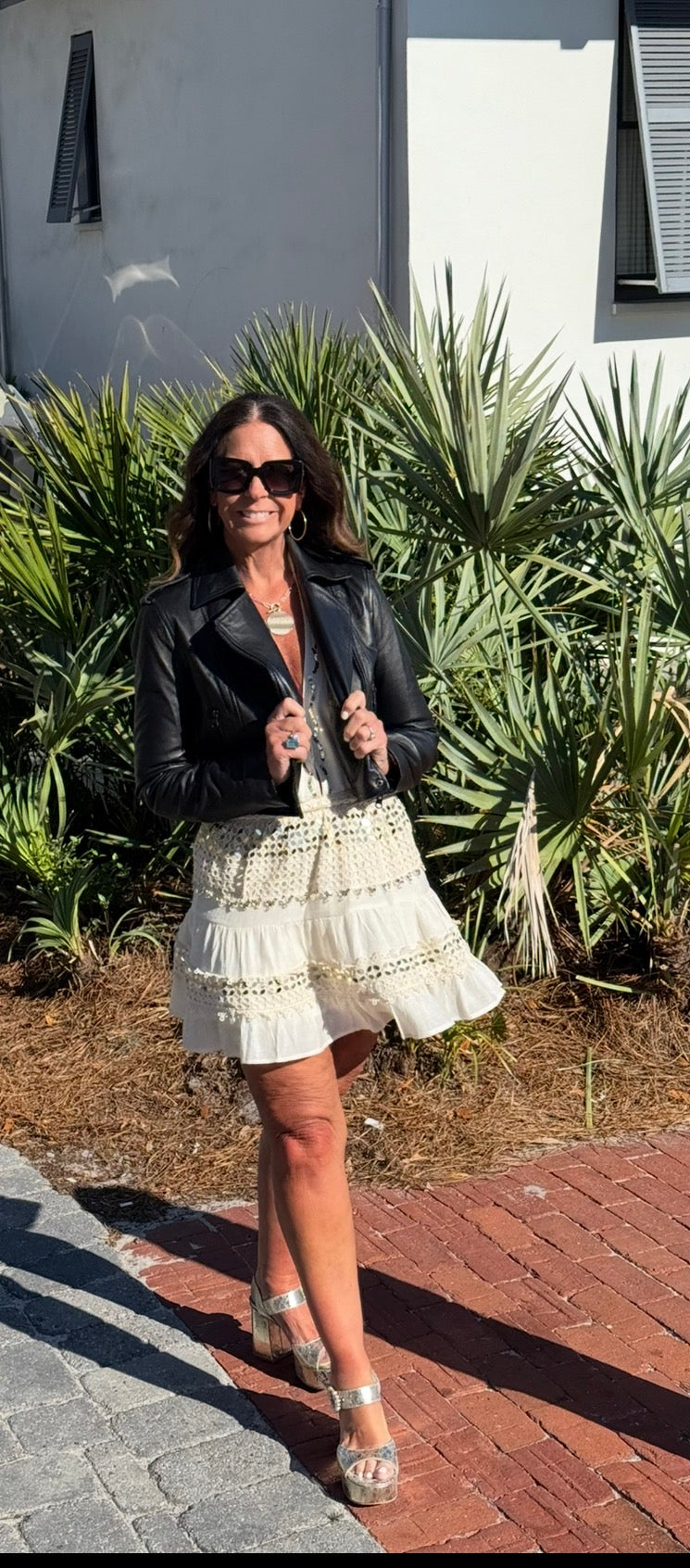 Woman in a black leather jacket and white lace dress standing outdoors with plants in the background