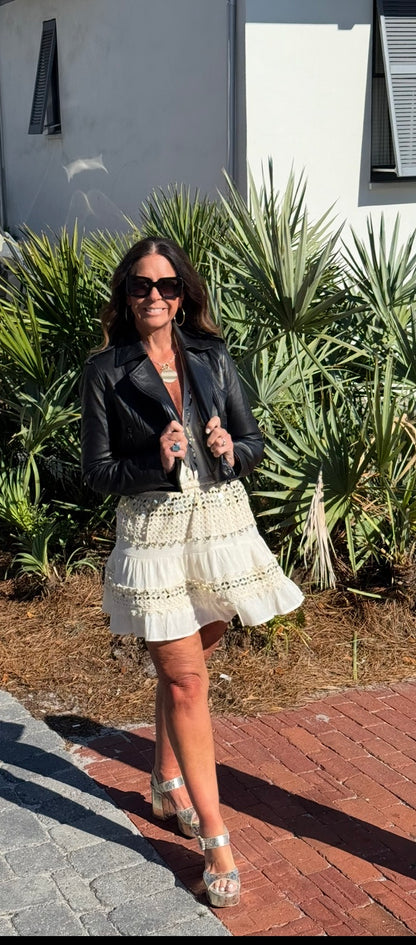 Woman in a black leather jacket and white lace dress standing outdoors with plants in the background