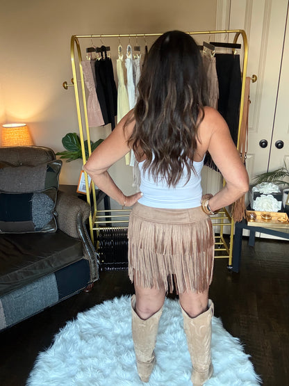 Woman modeling a tan fringe mini skirt in a home boutique.