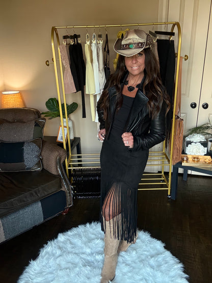 Woman in a stylish outfit with a black leather jacket and fringe dress, standing in a room with a clothing rack and furniture.