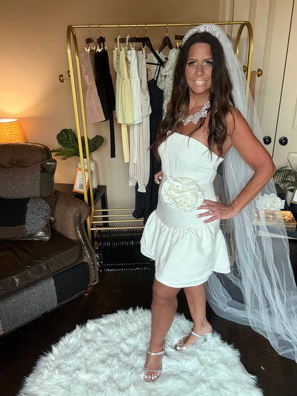 Woman in a white dress with a veil and bouquet standing in a room with furniture and decor.