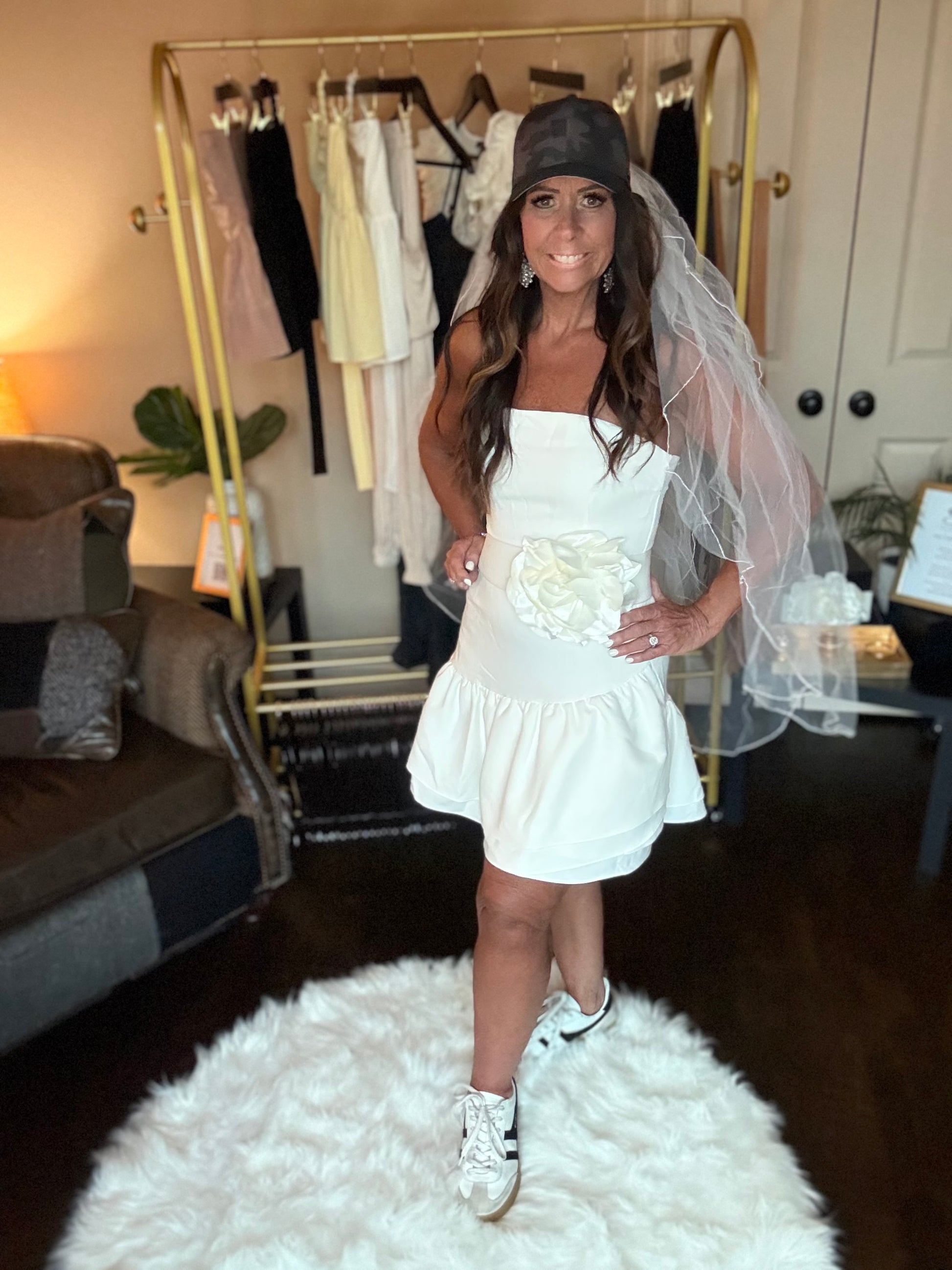 Woman in a white dress with a veil and capris standing on a white rug in a room with a clothing rack.